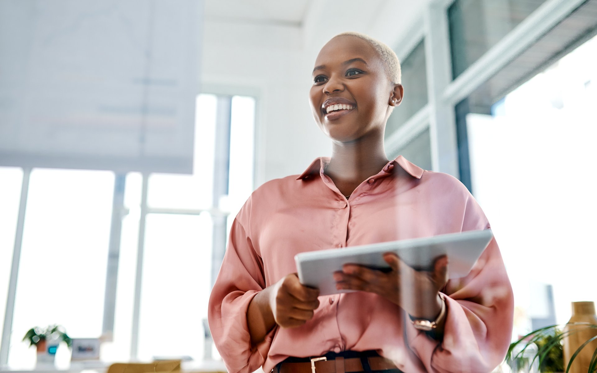 Smiling young woman in a pink shirt holding a tablet, standing in a bright office environment.