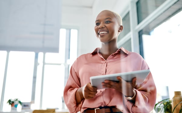 Smiling young woman in a pink shirt holding a tablet, standing in a bright office environment.
