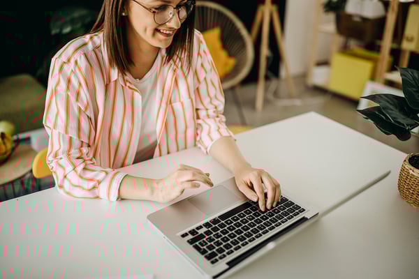 En kvinna med glasögon och en randig skjorta sitter vid ett skrivbord och arbetar på en laptop, leende och fokuserad.