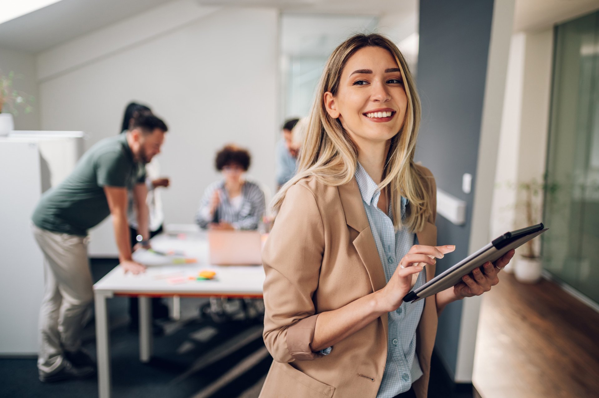 A blonde woman in a beige blazer and blue shirt smiles while holding a tablet in a modern office space.