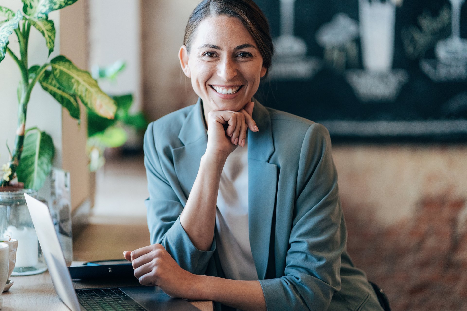 Smiling woman in a blazer sitting at a desk with a laptop in front of her.