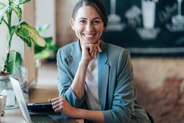 Smiling woman in a blazer sitting at a desk with a laptop in front of her.