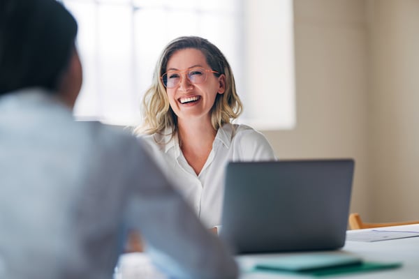 Smiling woman in front of a laptop, sitting across from another person seen from behind