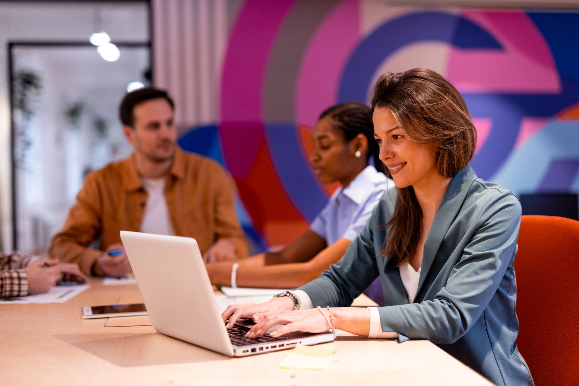Kvinna arbetar vid en laptop i ett färgglatt, modernt kontorslandskap med två kollegor i bakgrunden.