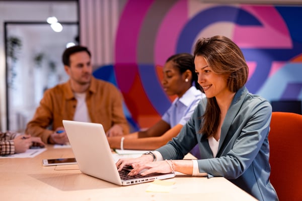 Kvinna arbetar vid en laptop i ett färgglatt, modernt kontorslandskap med två kollegor i bakgrunden.