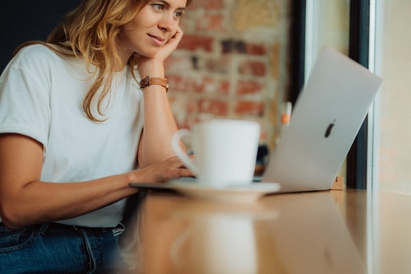 Young woman sitting at a table with a coffee cup and laptop, her head resting on her hand.