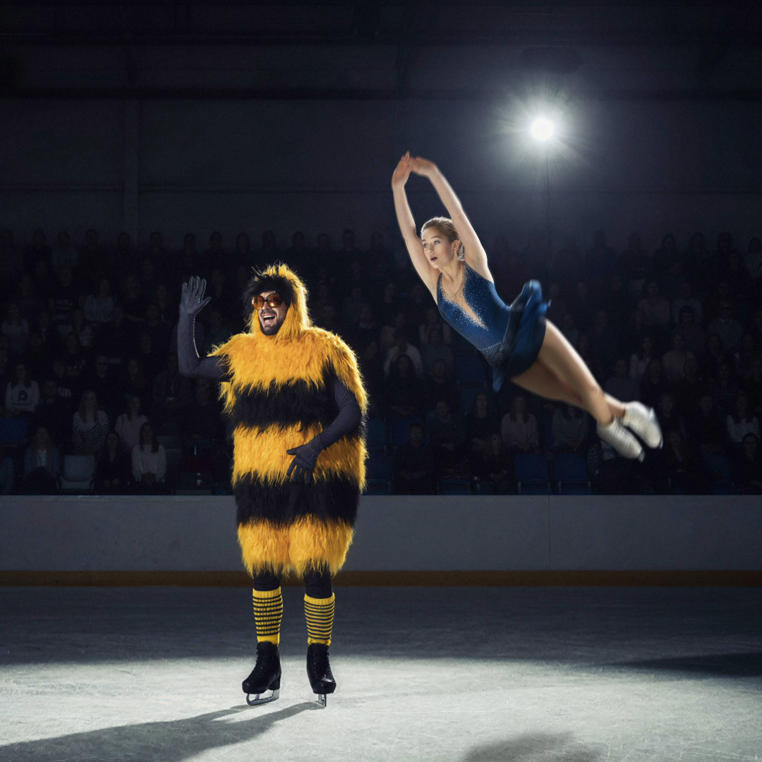 A figure skater in a blue dress performs a high jump in the air on an ice rink. Next to her stands a waving mascot dressed as a large, furry bumblebee wearing sunglasses. In the background, a dark arena stand with an audience is visible.
