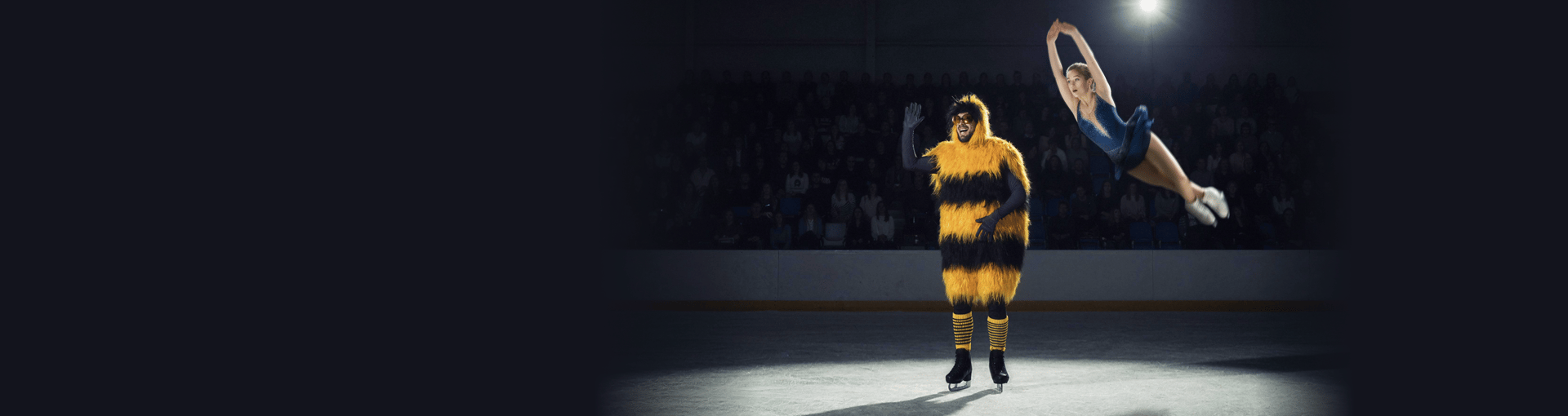 A figure skater in a blue dress performs a high jump in the air on an ice rink. Next to her stands a waving mascot dressed as a large, furry bumblebee wearing sunglasses. In the background, a dark arena stand with an audience is visible.